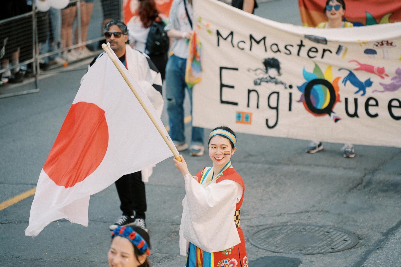 A participant in traditional attire waves a Japanese flag at a parade, with a 'McMaster EngiQueers' banner in the background.
