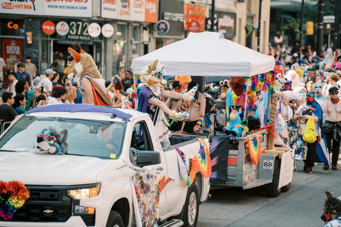 A vibrant parade scene with participants in colorful costumes and animal masks, riding a decorated float and white pickup truck, surrounded by a lively crowd.