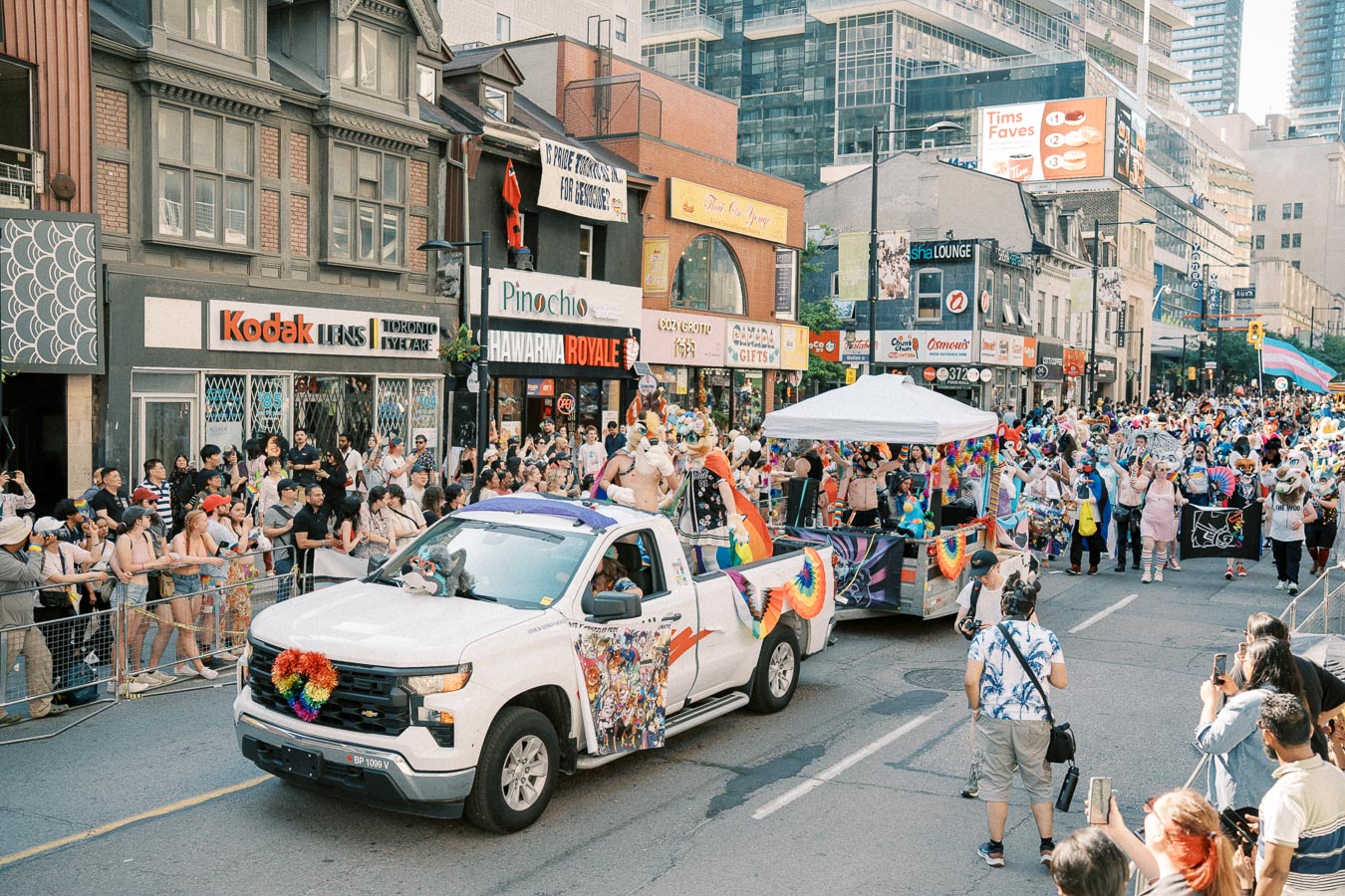 A lively parade scene featuring a decorated white truck and participants in colorful costumes, with spectators lining the street in an urban setting.
