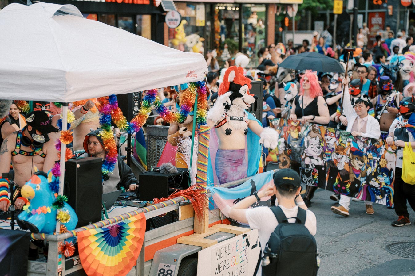 A vibrant parade scene showcasing participants in colorful costumes and furry outfits, celebrating with rainbow decorations and a sign that reads 'We're here, we're queer.'
