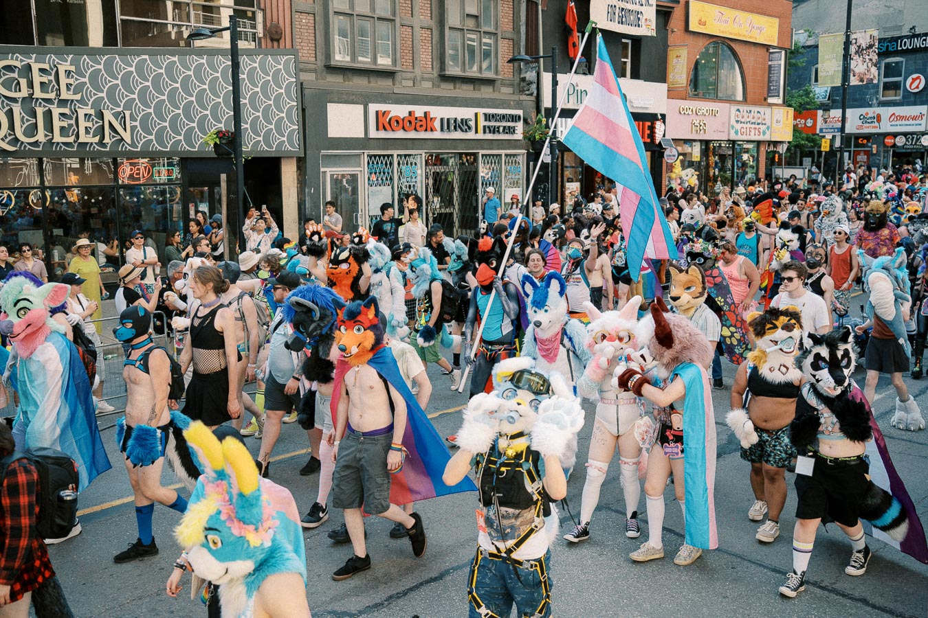 A vibrant parade with participants dressed in colorful animal costumes, celebrating diversity and inclusion. A transgender pride flag is prominently displayed, adding to the festive atmosphere.