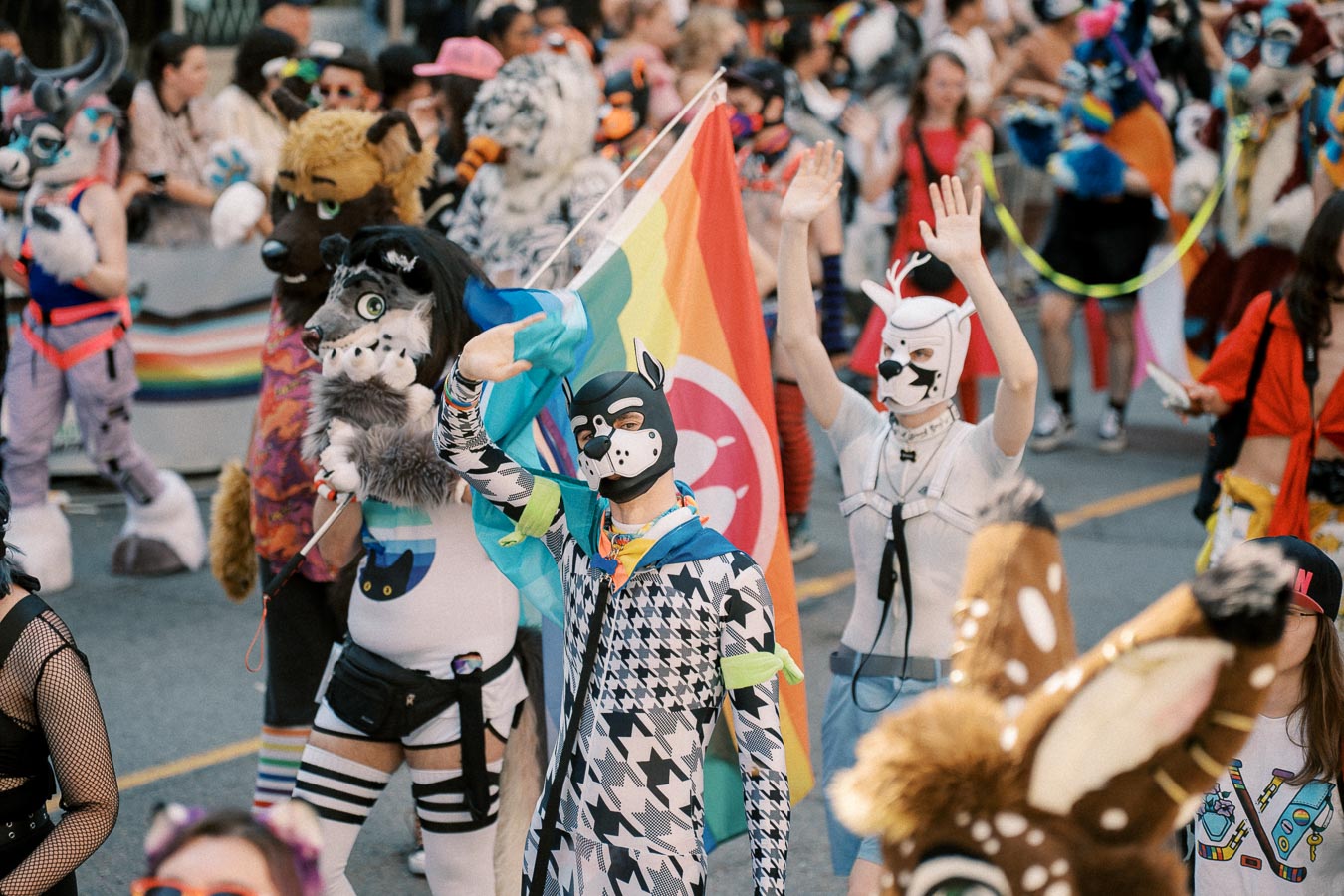 Participants in colorful animal costumes march in a parade, waving a rainbow flag.