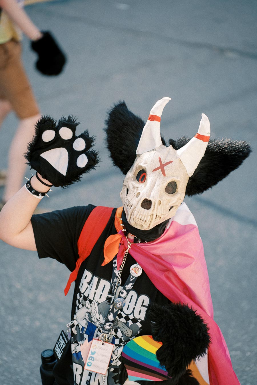 A person dressed in a creative costume featuring a skull mask with horns and large ears, complemented by a paw glove and a colorful cape, participates in a public event.