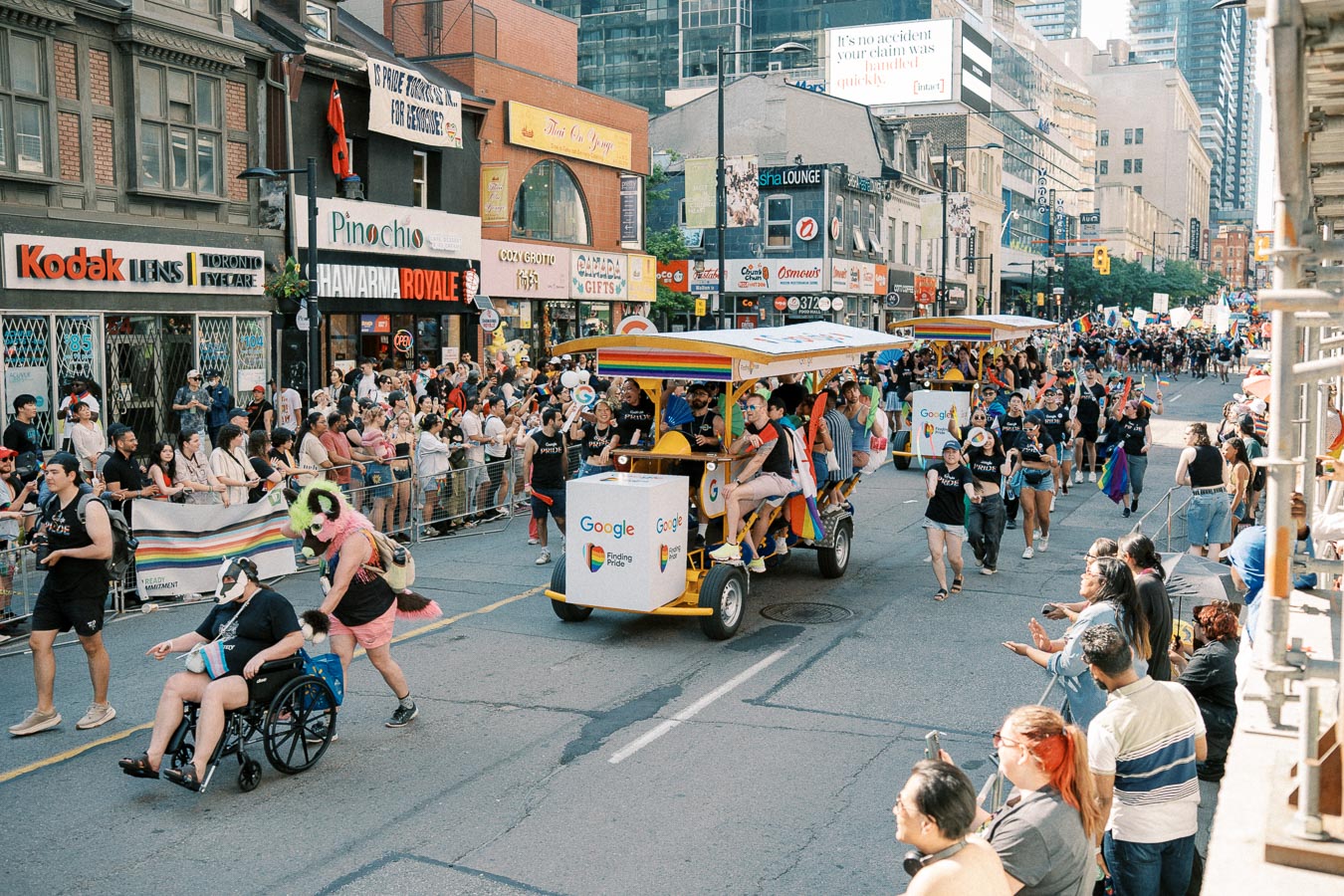 A lively parade scene featuring a Google-branded vehicle and participants in colorful outfits, with spectators lining the street.