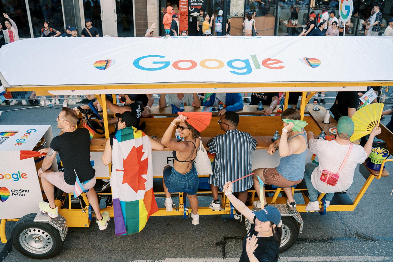 Participants ride a Google-branded pedal-powered vehicle at a pride parade, celebrating with rainbow flags and fans.