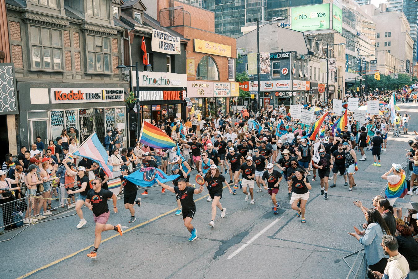 Participants in a vibrant parade march down a city street, waving rainbow flags and banners, while spectators cheer from the sidelines.