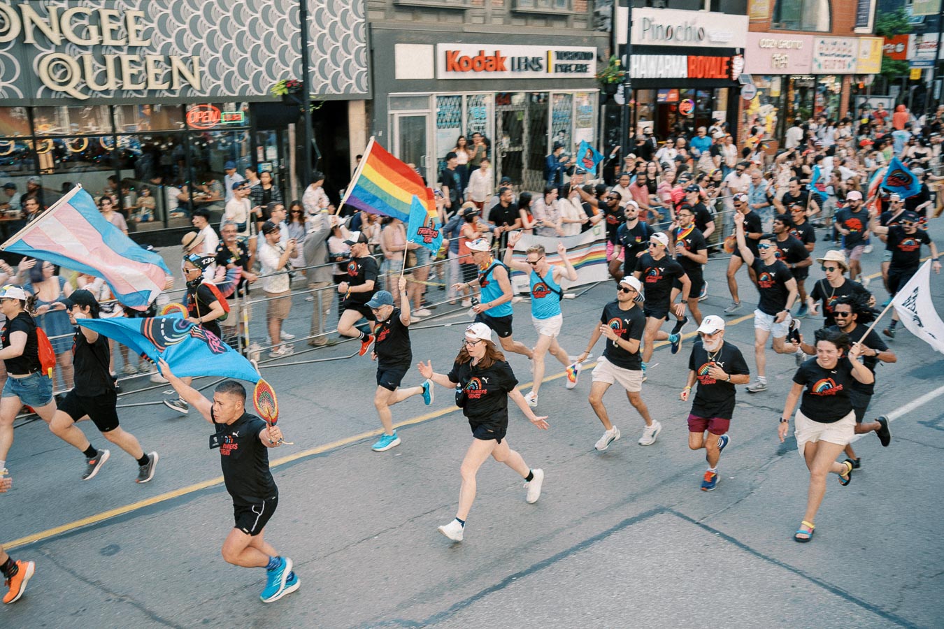 Participants run enthusiastically in a pride parade, waving rainbow and transgender flags, as onlookers cheer from the sidelines.