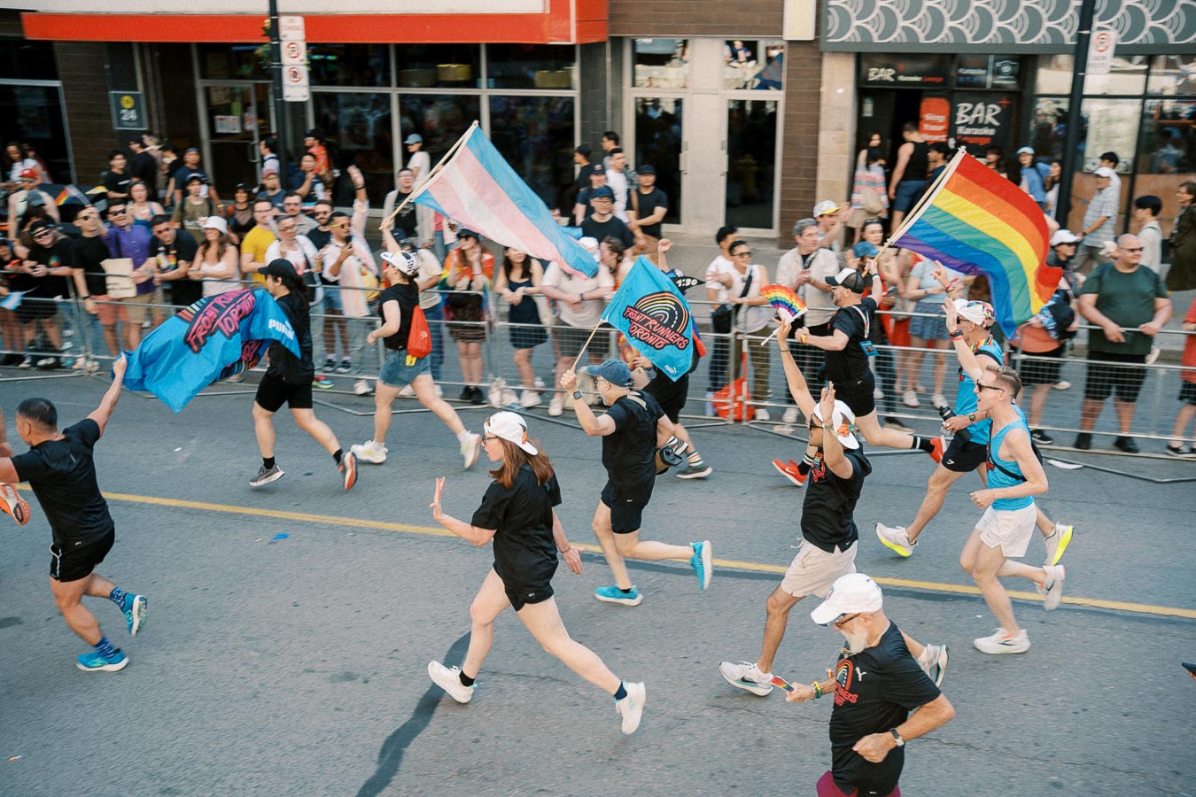 Participants run in a pride parade, waving rainbow and transgender flags, as spectators cheer from the sidelines.