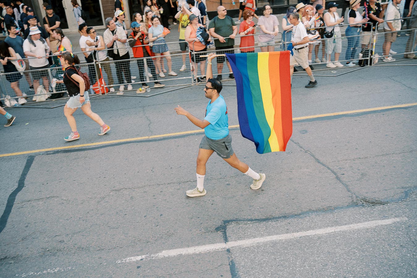 A participant carries a rainbow flag during a parade, with spectators watching from behind a barrier.
