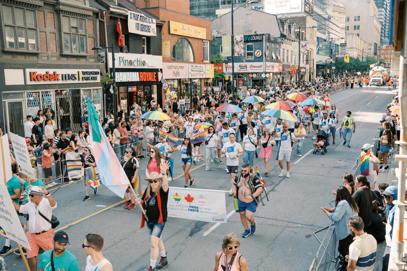 A lively street parade featuring participants with rainbow umbrellas and flags, celebrating diversity and inclusion. Spectators line the street, enjoying the festive atmosphere.