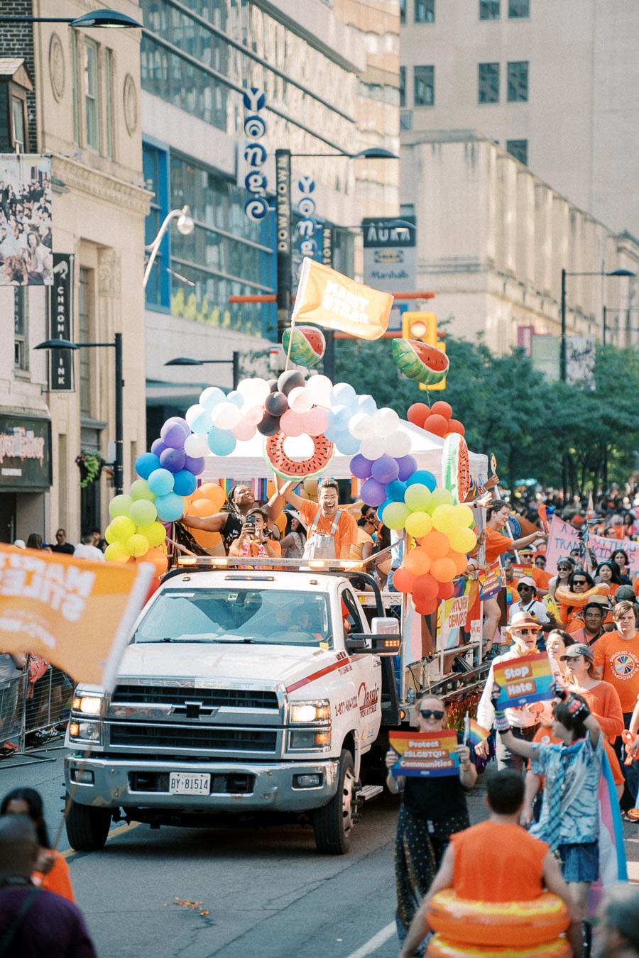 A vibrant pride parade featuring a float adorned with colorful balloons and participants in orange shirts, celebrating diversity and inclusion on a city street.