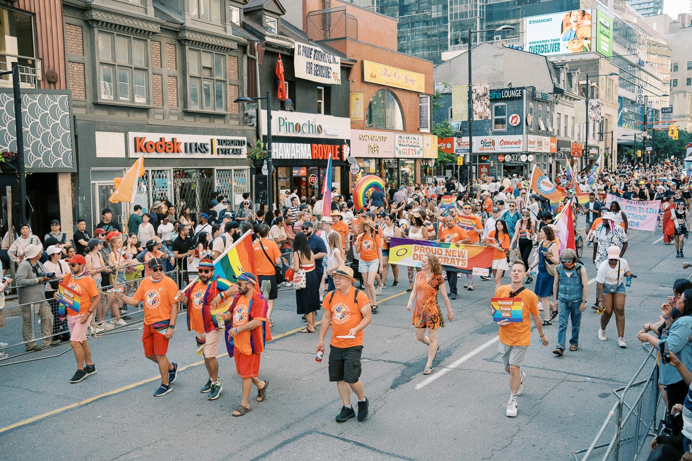 Participants in a colorful parade march down a city street, celebrating with rainbow flags and banners, while spectators watch from the sidelines.