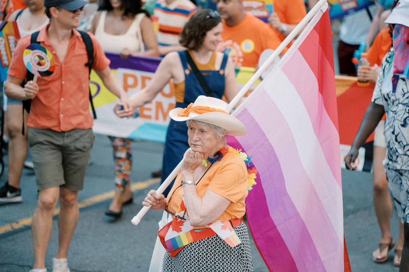An elderly woman participates in a pride parade, holding a pride flag and wearing a colorful outfit, surrounded by other parade-goers.