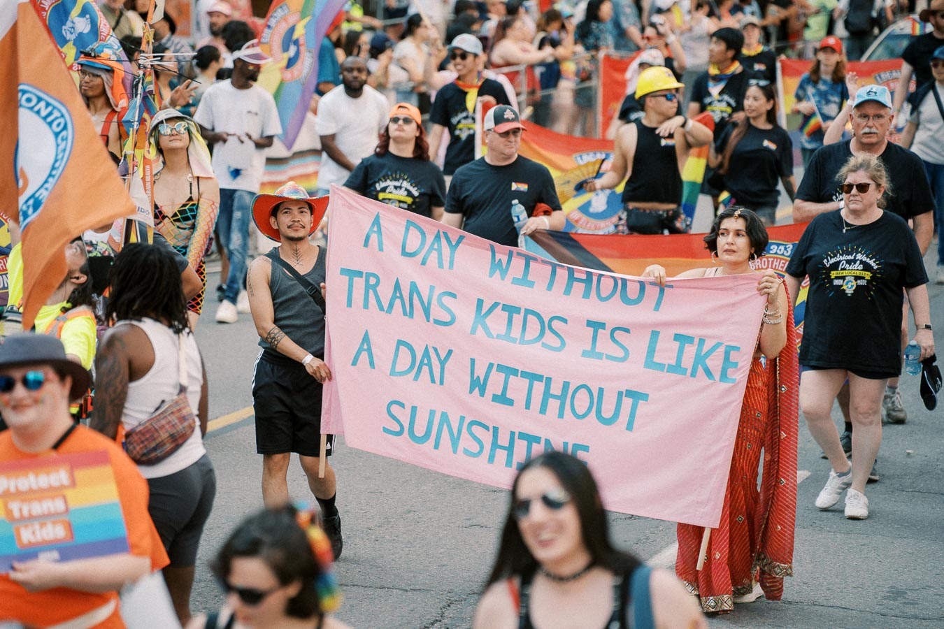 Participants in a pride parade hold a banner reading 'A Day Without Trans Kids is Like a Day Without Sunshine,' advocating for transgender rights.