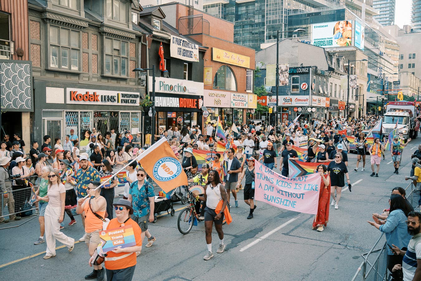 Participants in a lively parade march down a city street, displaying rainbow flags and banners advocating for LGBTQ+ rights, while spectators line the sidewalks.