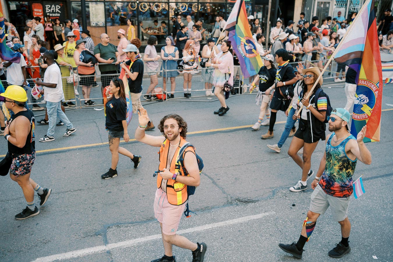 Participants in a pride parade march down the street, waving rainbow flags and wearing vibrant attire. Spectators line the sidewalk, watching the procession.
