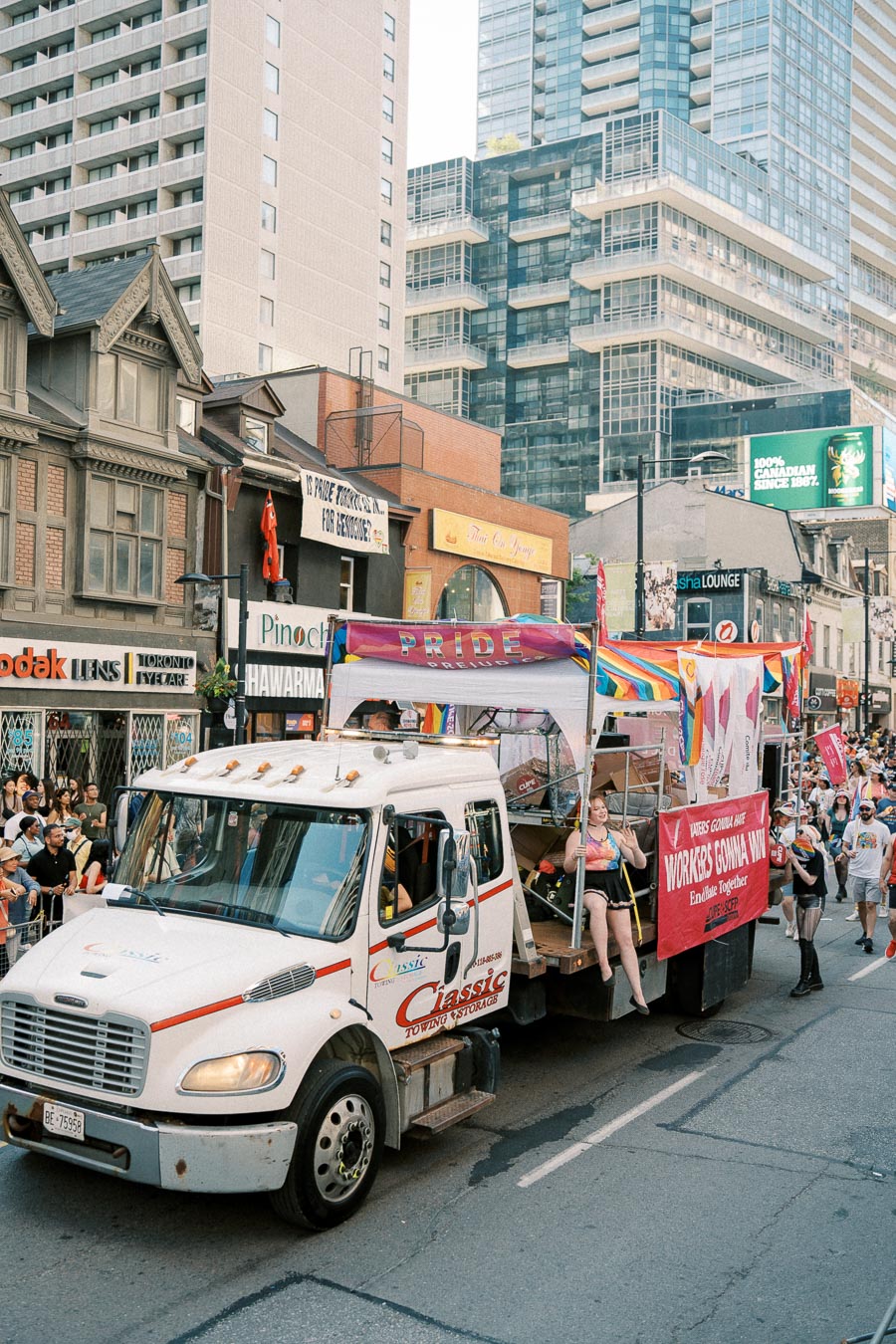 A parade float featuring a white truck with people celebrating, adorned with pride flags and banners, moves through a city street lined with spectators and tall buildings.
