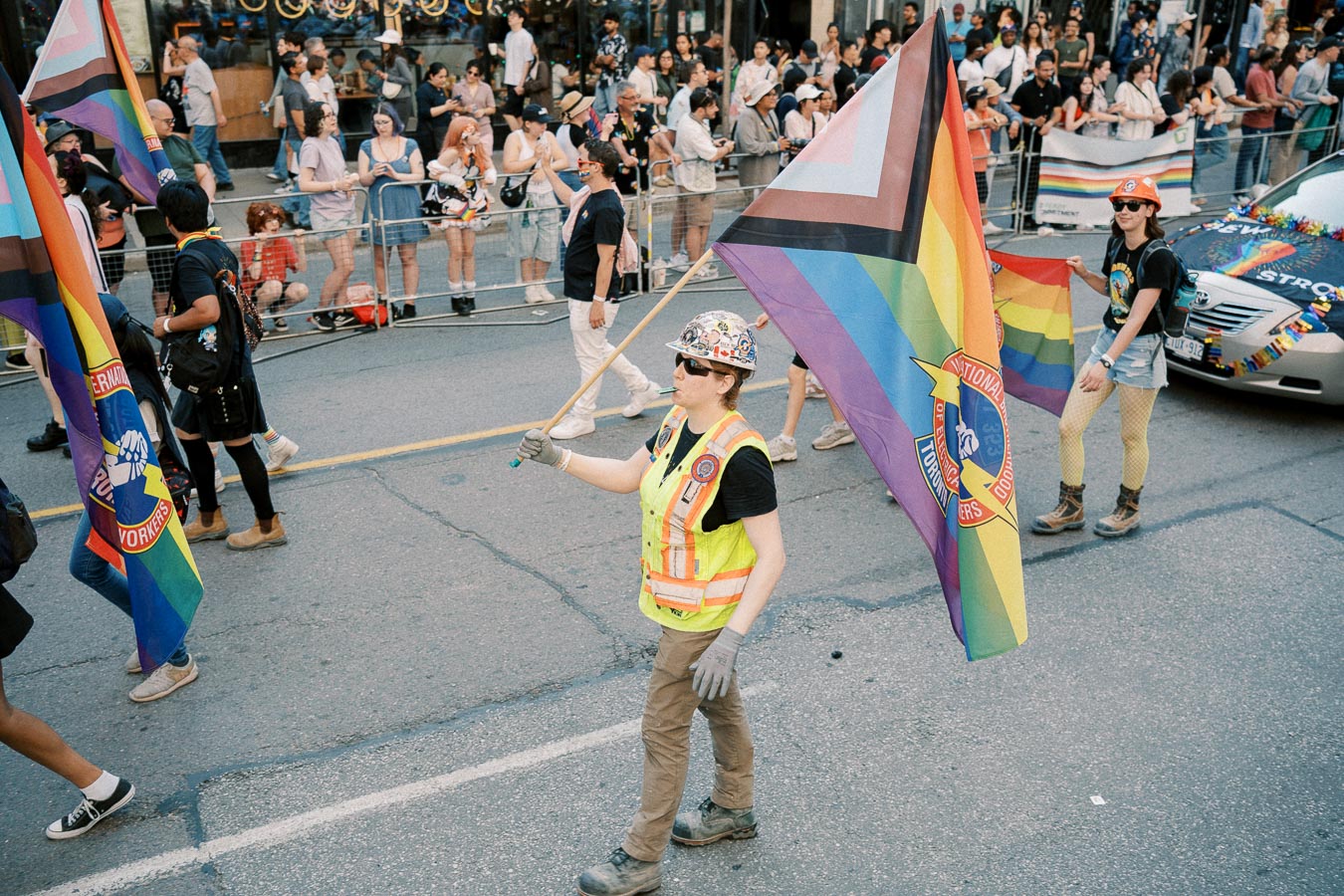 Participants in a pride parade march down the street holding rainbow flags, with onlookers gathered behind barriers.