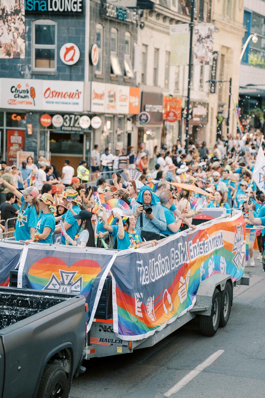 A colorful parade float with participants in blue shirts and rainbow-themed accessories, celebrating on a bustling city street.