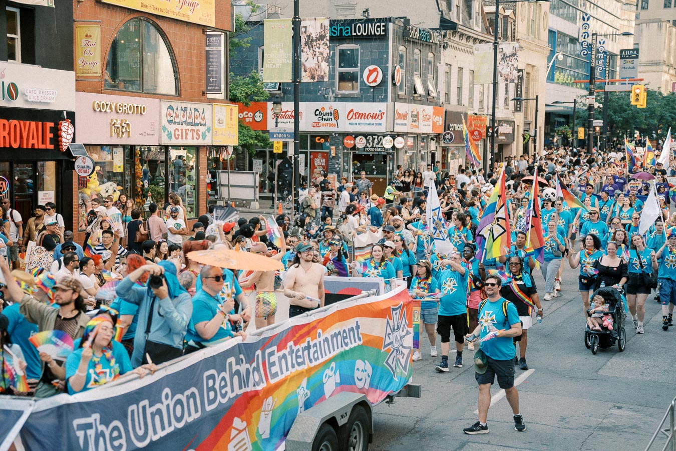Participants in a colorful parade march down a bustling street, celebrating with rainbow flags and vibrant attire.