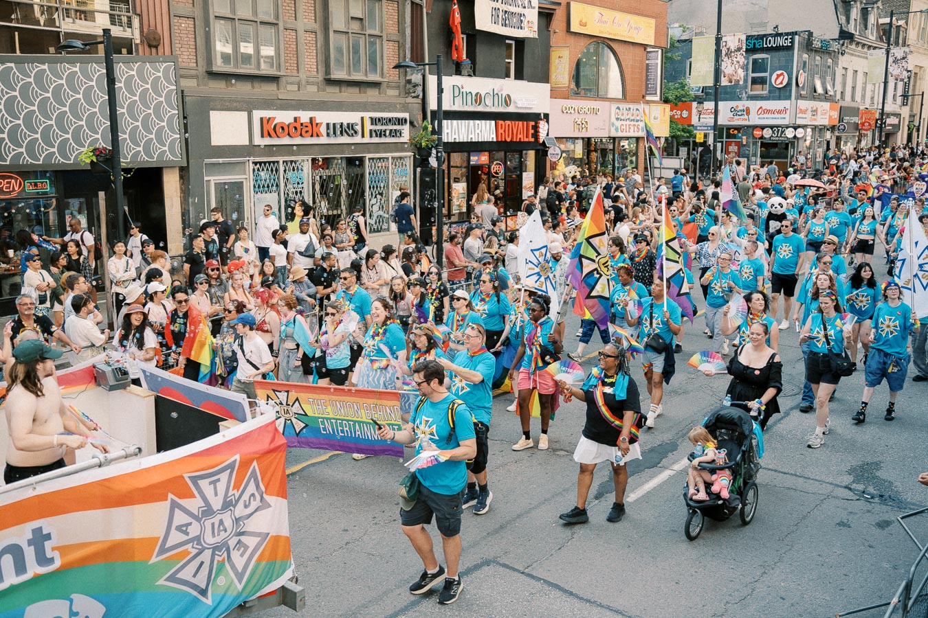 Participants in a colorful parade march down a city street, celebrating with rainbow flags and banners, surrounded by cheering spectators.