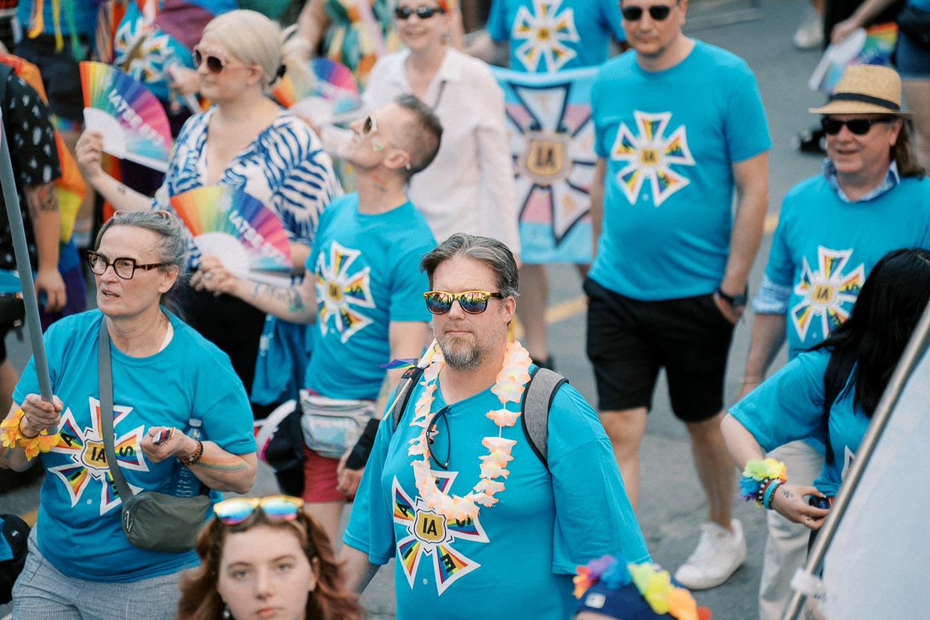 Participants in a parade wearing blue shirts with a colorful emblem, holding rainbow fans and adorned with vibrant accessories.