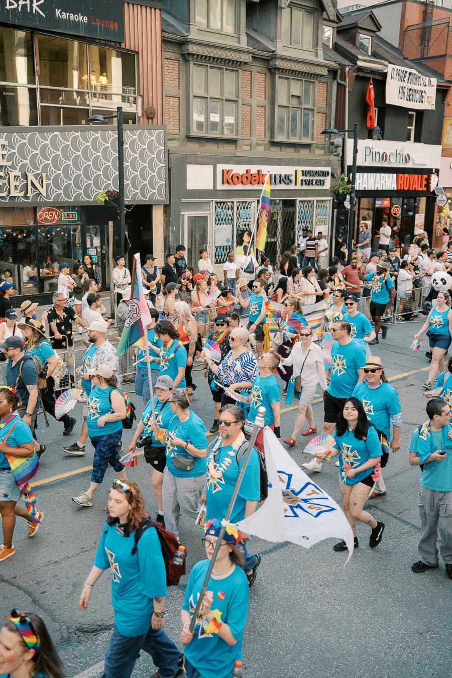 Participants in a pride parade march down a city street, wearing matching blue shirts and carrying flags, with spectators and storefronts lining the route.