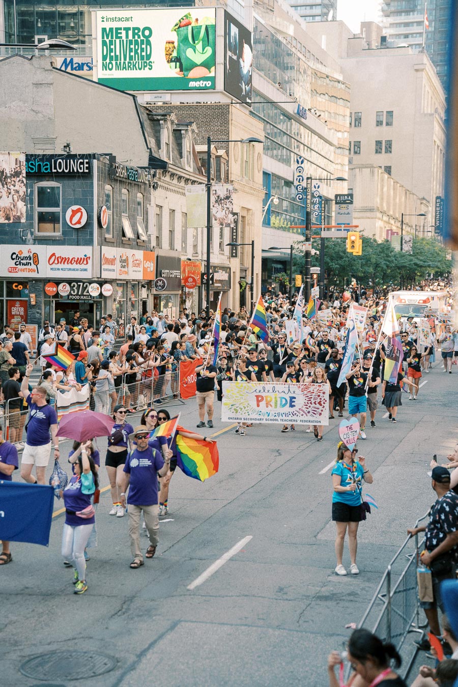Participants in a Pride parade march down a city street, waving rainbow flags and carrying banners, while spectators line the sidewalks.