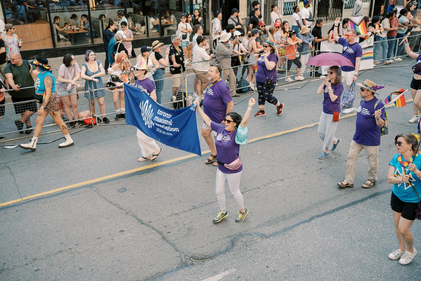 Participants from the Toronto Musicians Association march in a parade, showcasing vibrant attire and carrying a banner. Spectators line the street, enjoying the festive atmosphere.