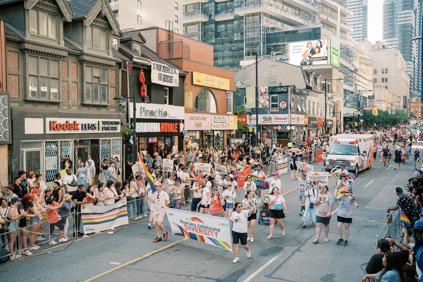 A lively parade celebrating diversity, featuring nurses marching with a banner, takes place on a bustling city street lined with spectators and various shops.