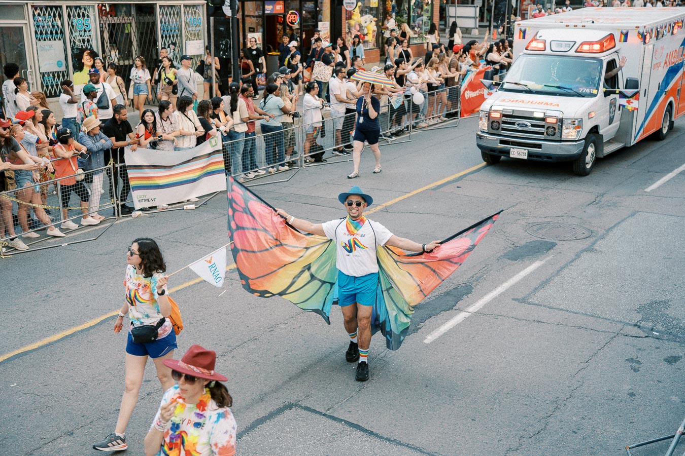 A vibrant scene from a pride parade featuring a participant in a rainbow butterfly costume, with an ambulance and cheering spectators lining the street.