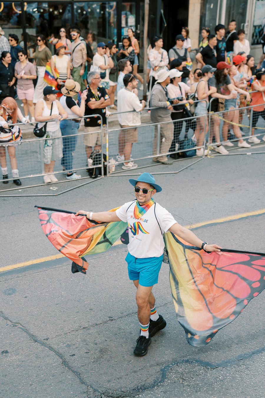 A participant in a pride parade wearing colorful butterfly wings and a rainbow-themed outfit, walking past a crowd of spectators.