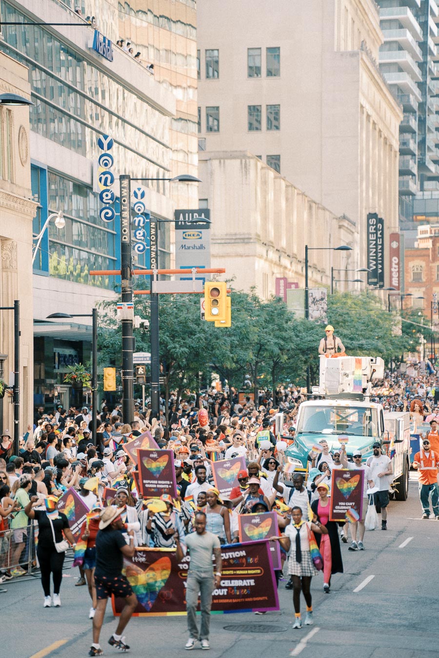 A lively parade featuring participants with rainbow-themed signs and banners, marching through a bustling city street.