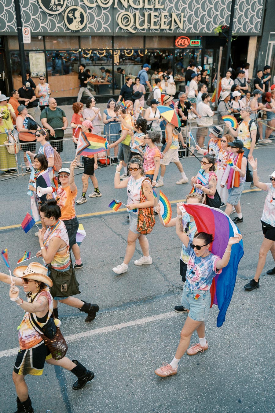 Participants in a pride parade march joyfully, waving rainbow flags and wearing vibrant attire, as onlookers cheer from the sidelines.