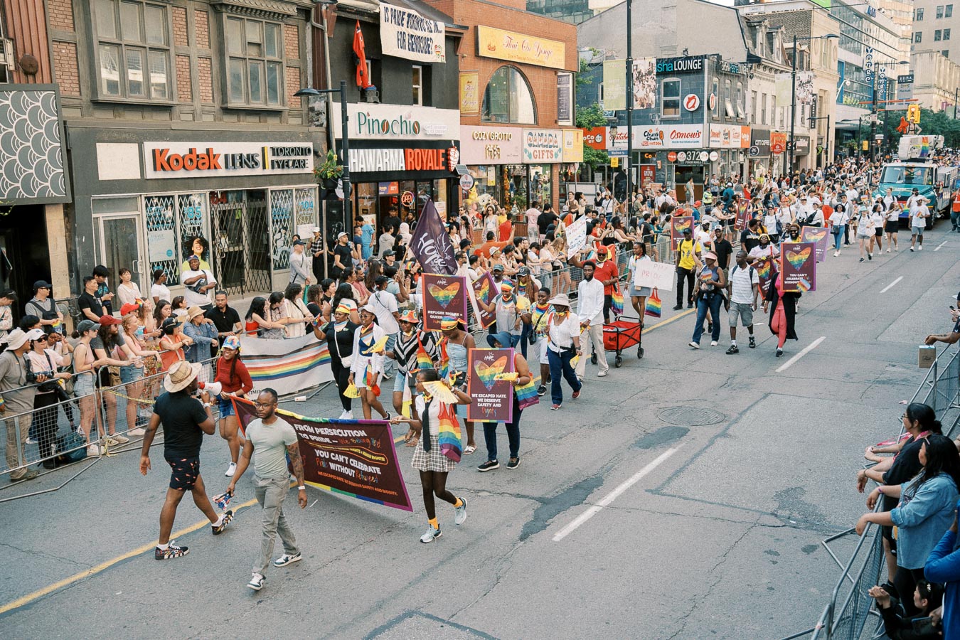 Participants in a pride parade march down a city street, holding colorful banners and rainbow flags, while spectators watch from the sidelines.