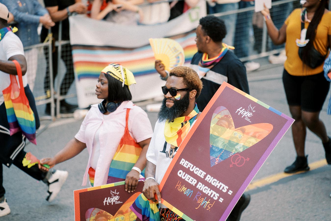 Participants in a pride parade hold signs and rainbow flags advocating for refugee and queer rights.
