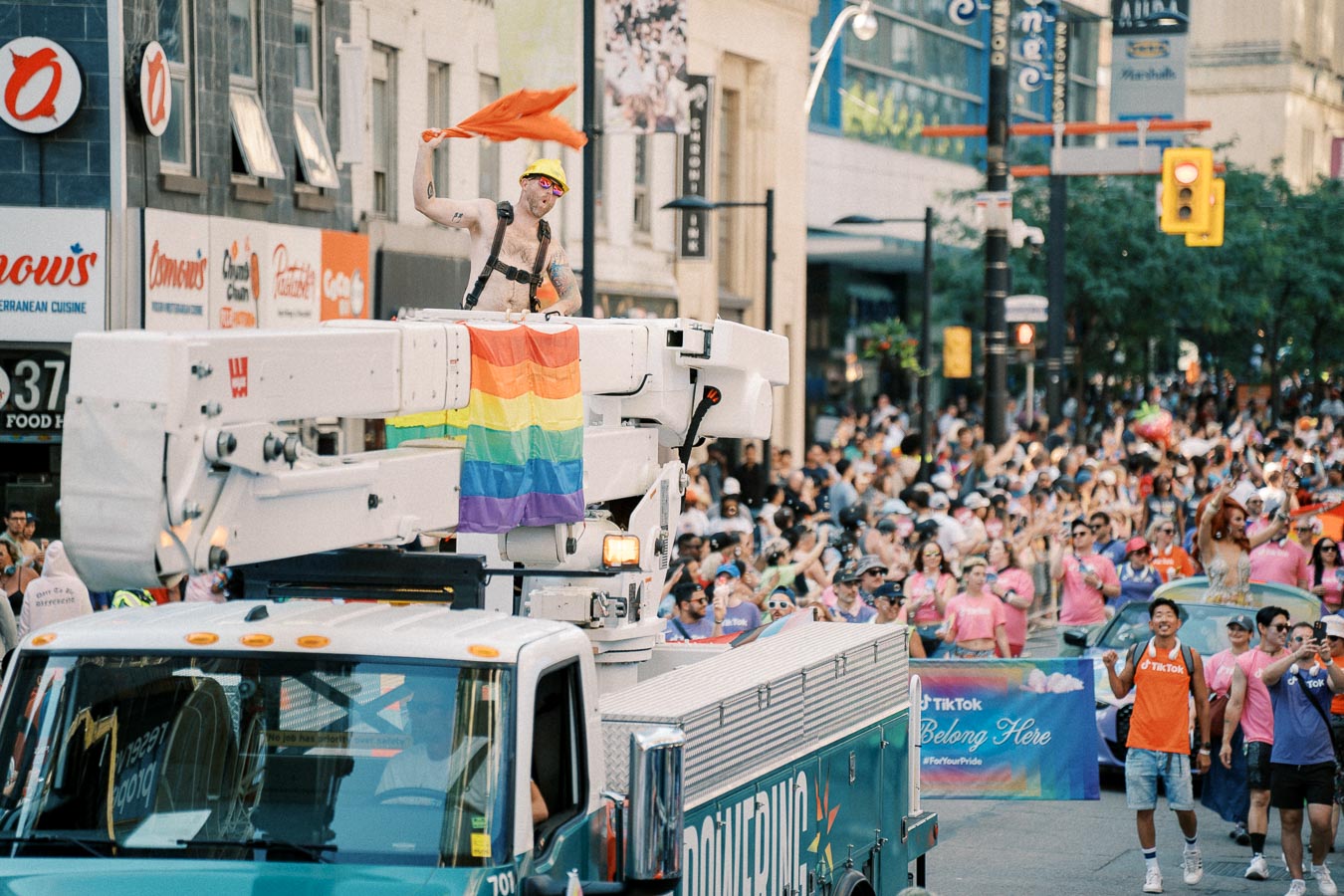 A lively pride parade scene featuring a person waving an orange flag from a utility truck adorned with a rainbow flag, surrounded by a cheering crowd.