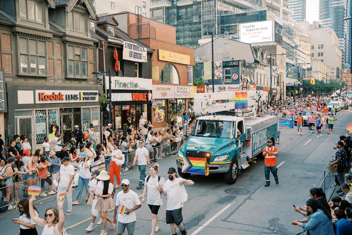 A lively parade celebrating diversity and inclusion, featuring participants with rainbow flags and a decorated truck, set against a backdrop of urban shops and buildings.