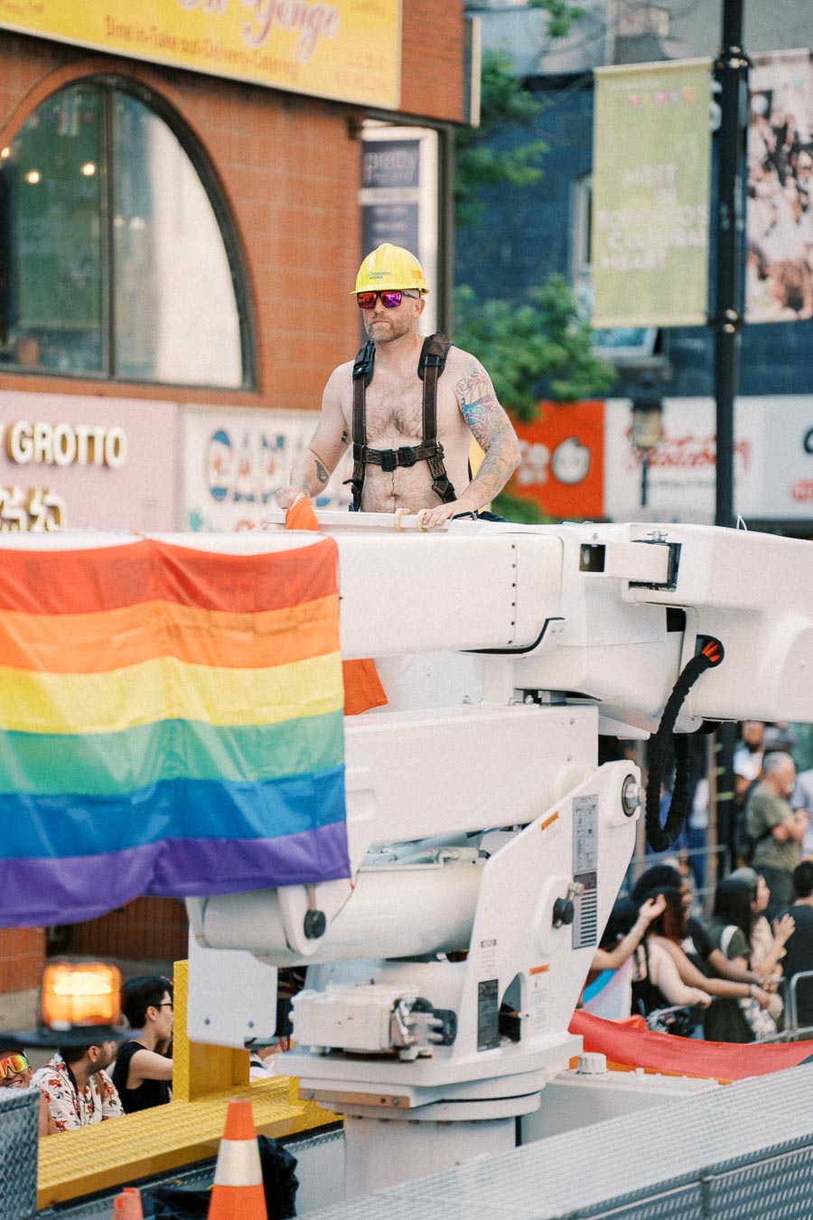 A person in a yellow hard hat and sunglasses stands on a platform adorned with a rainbow flag during a parade in an urban area.