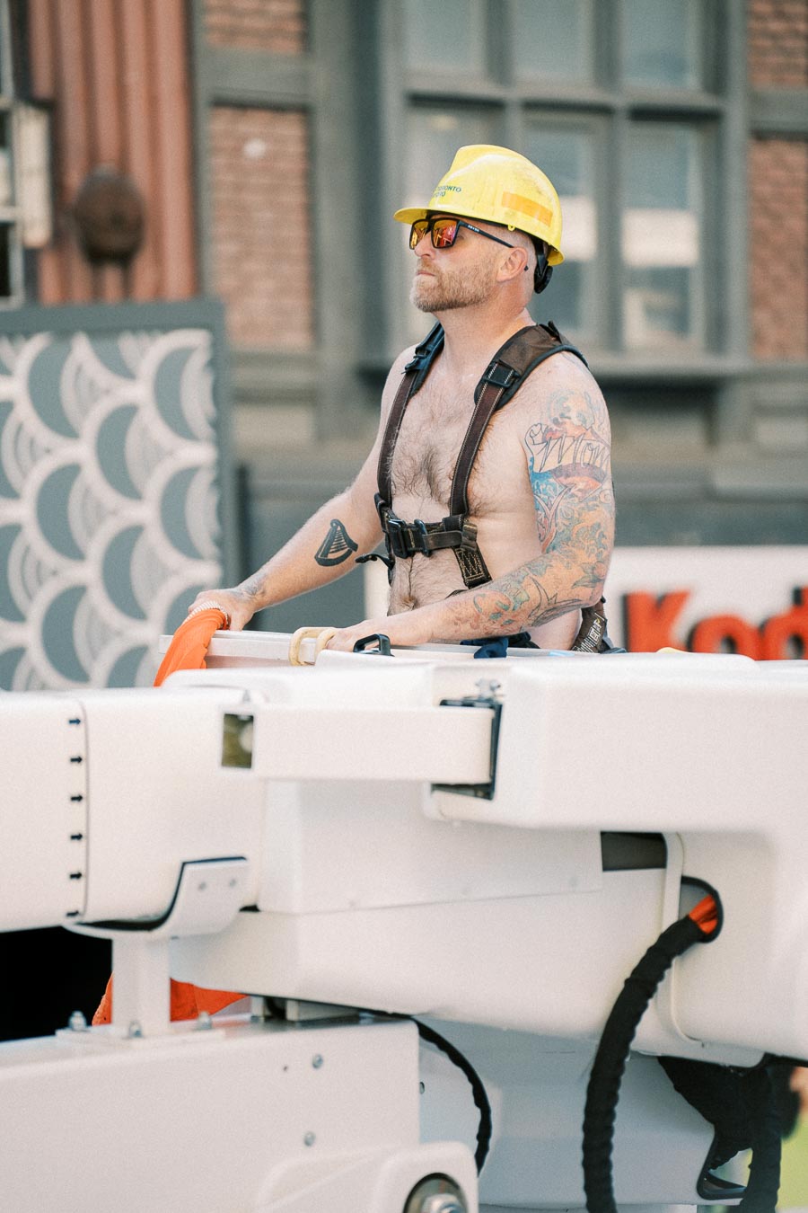 A construction worker, sporting a yellow hard hat and sunglasses, operates machinery on a city street. His arms are adorned with colorful tattoos, and he is wearing a safety harness.