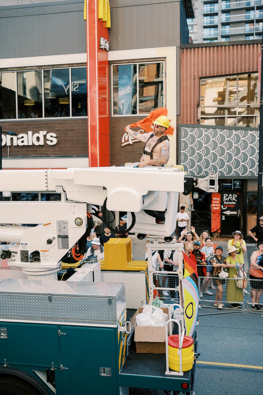 A person wearing a hard hat and harness stands on a lift truck during a parade, with a McDonald's restaurant in the background and a crowd of spectators.