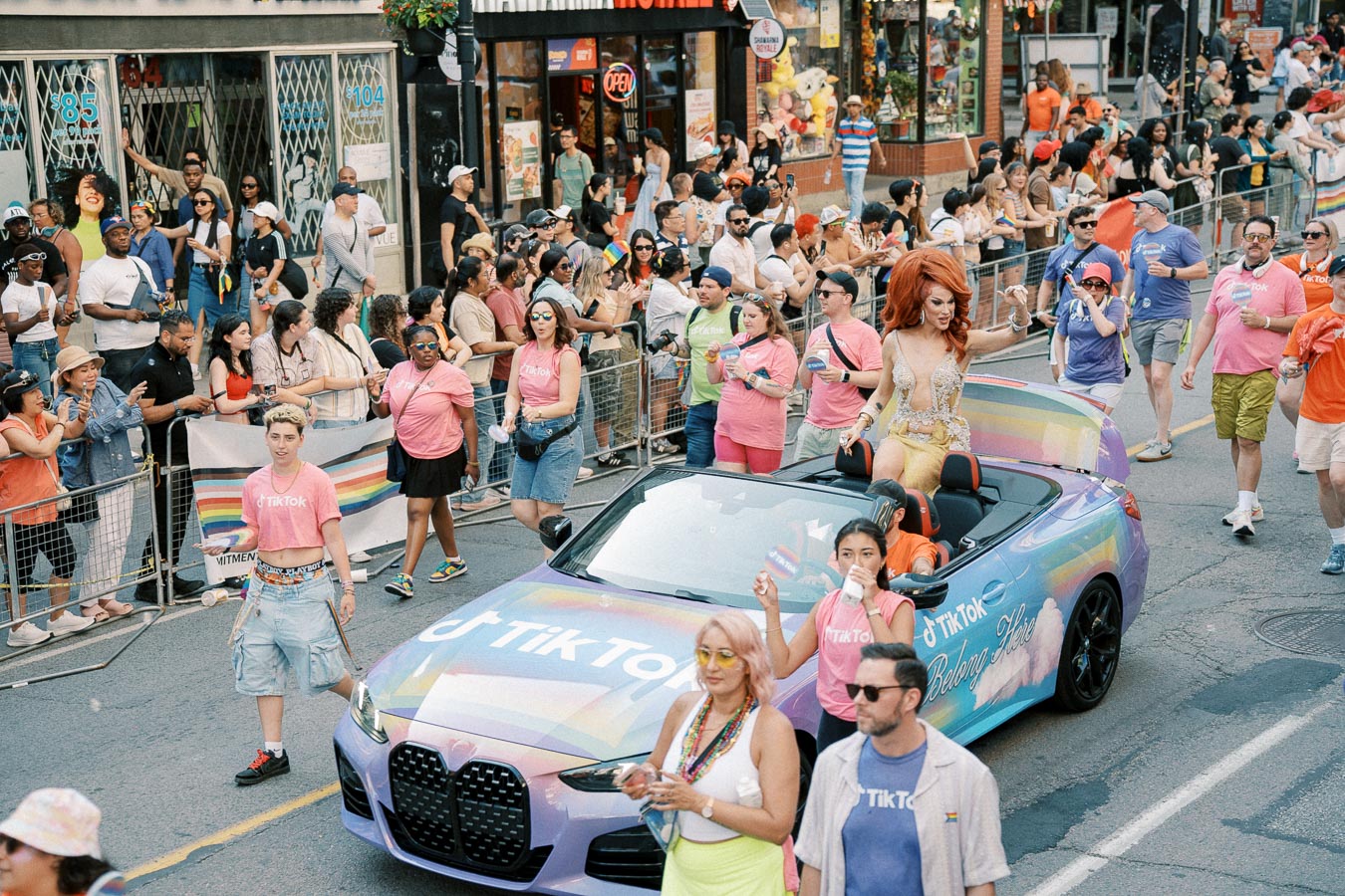 A vibrant parade scene featuring a TikTok-branded convertible with rainbow designs, surrounded by participants and spectators in colorful attire.