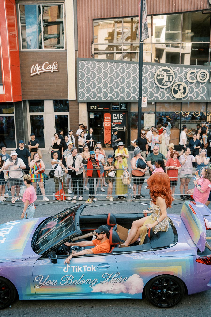 A vibrant parade scene featuring a TikTok-branded convertible with the message 'You Belong Here', accompanied by a glamorous participant and enthusiastic spectators.