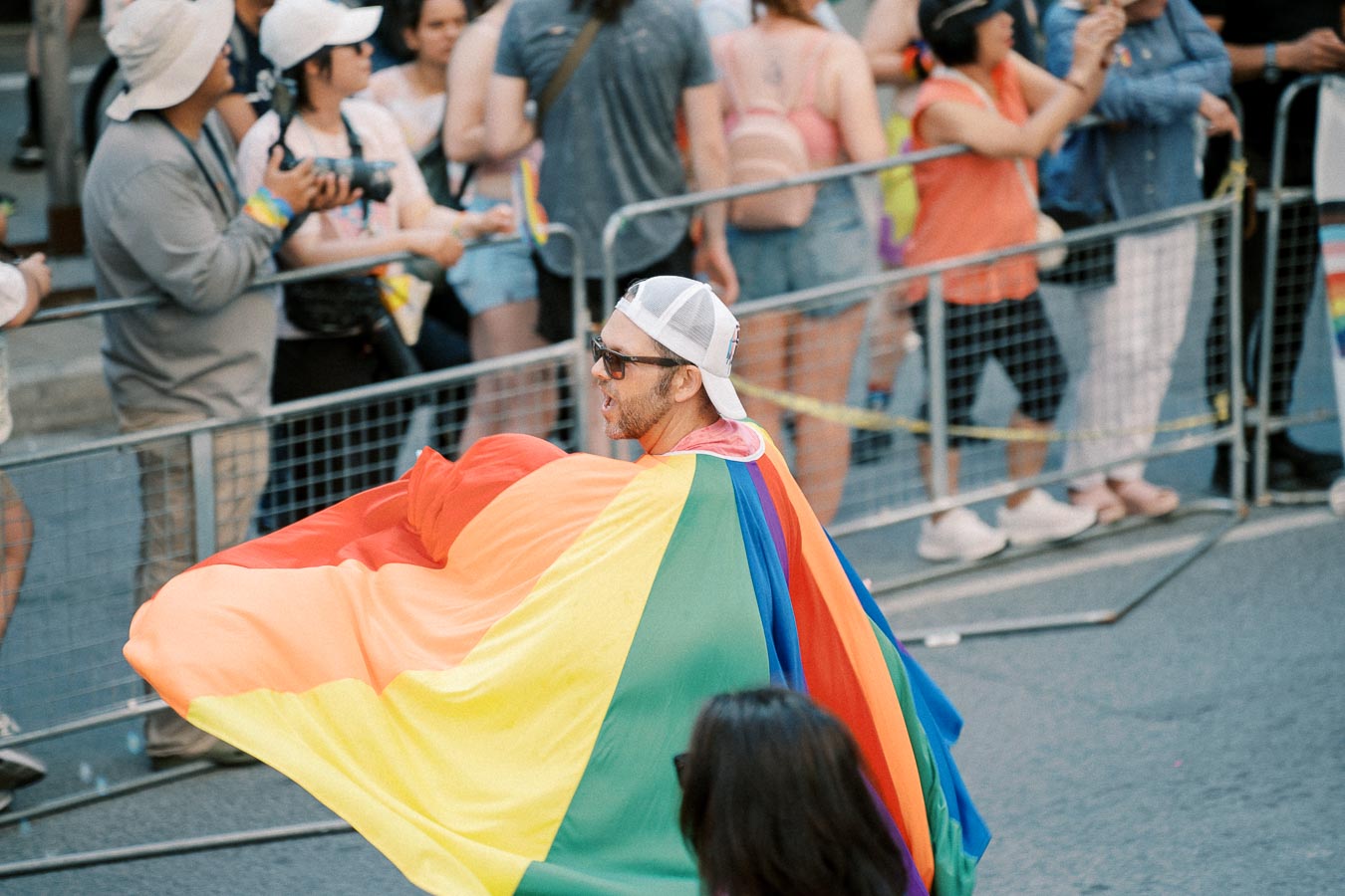 A participant draped in a rainbow flag walks in a pride parade, with onlookers capturing the moment.