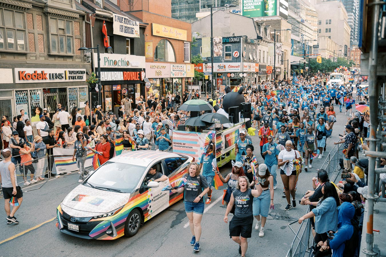A lively parade in Toronto featuring a rainbow-decorated car and a Centennial College float, with enthusiastic participants and spectators celebrating diversity and inclusion.