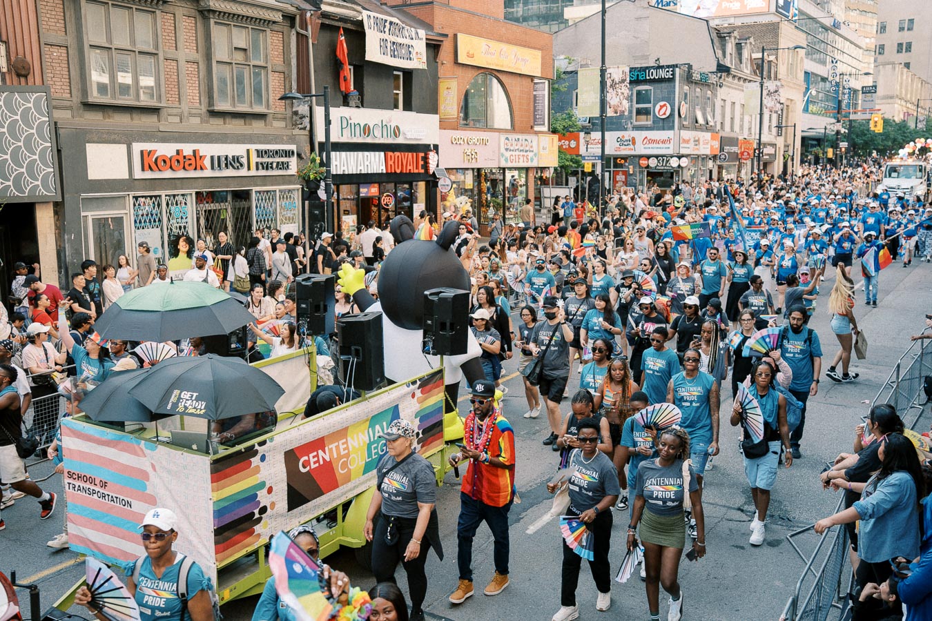 A lively parade featuring Centennial College participants celebrating Pride, with colorful shirts, flags, and a festive float. The street is bustling with spectators and lined with various shops.
