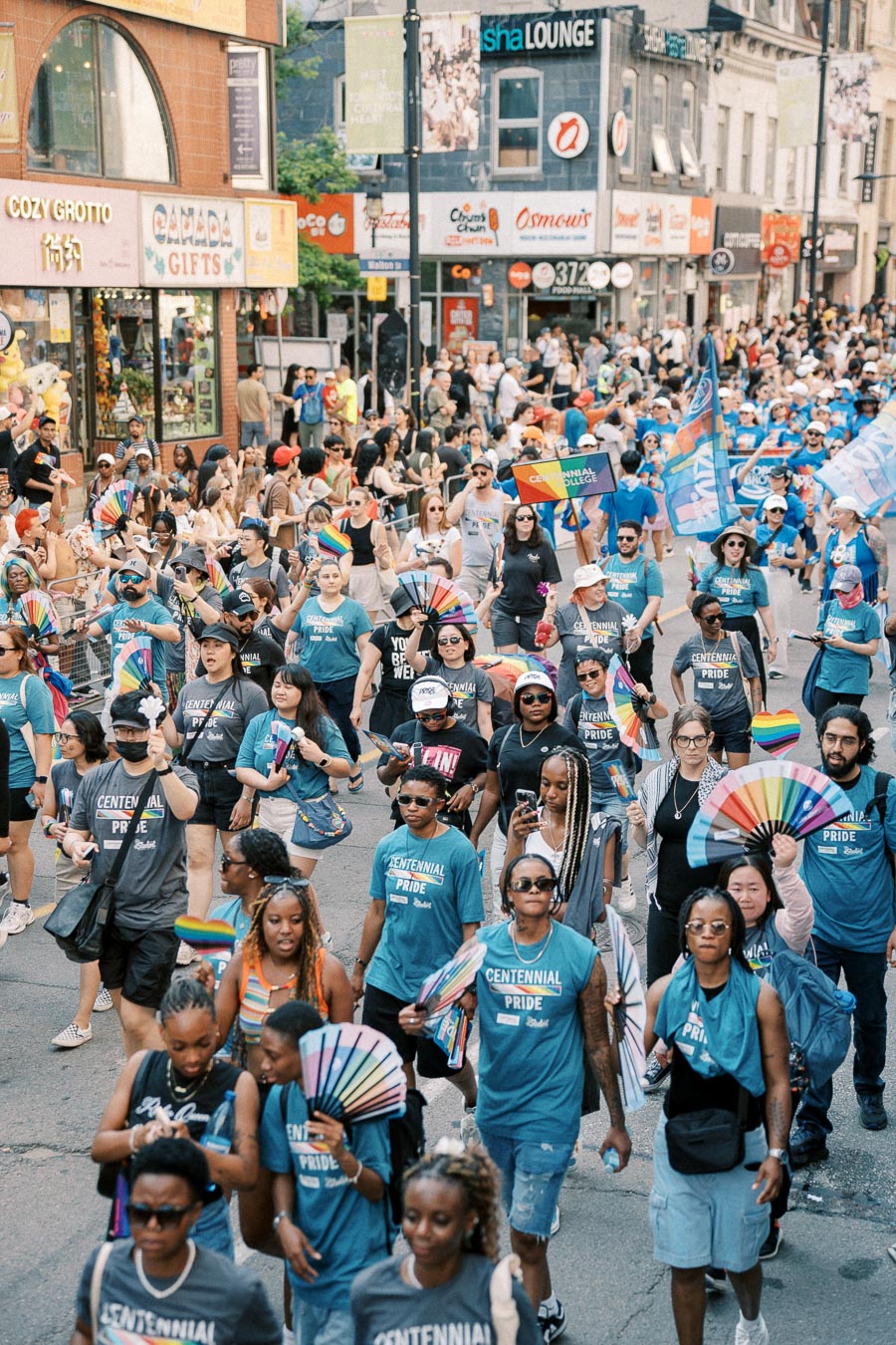 Participants in a pride parade wearing Centennial Pride shirts and holding rainbow flags and fans, marching down a bustling city street.