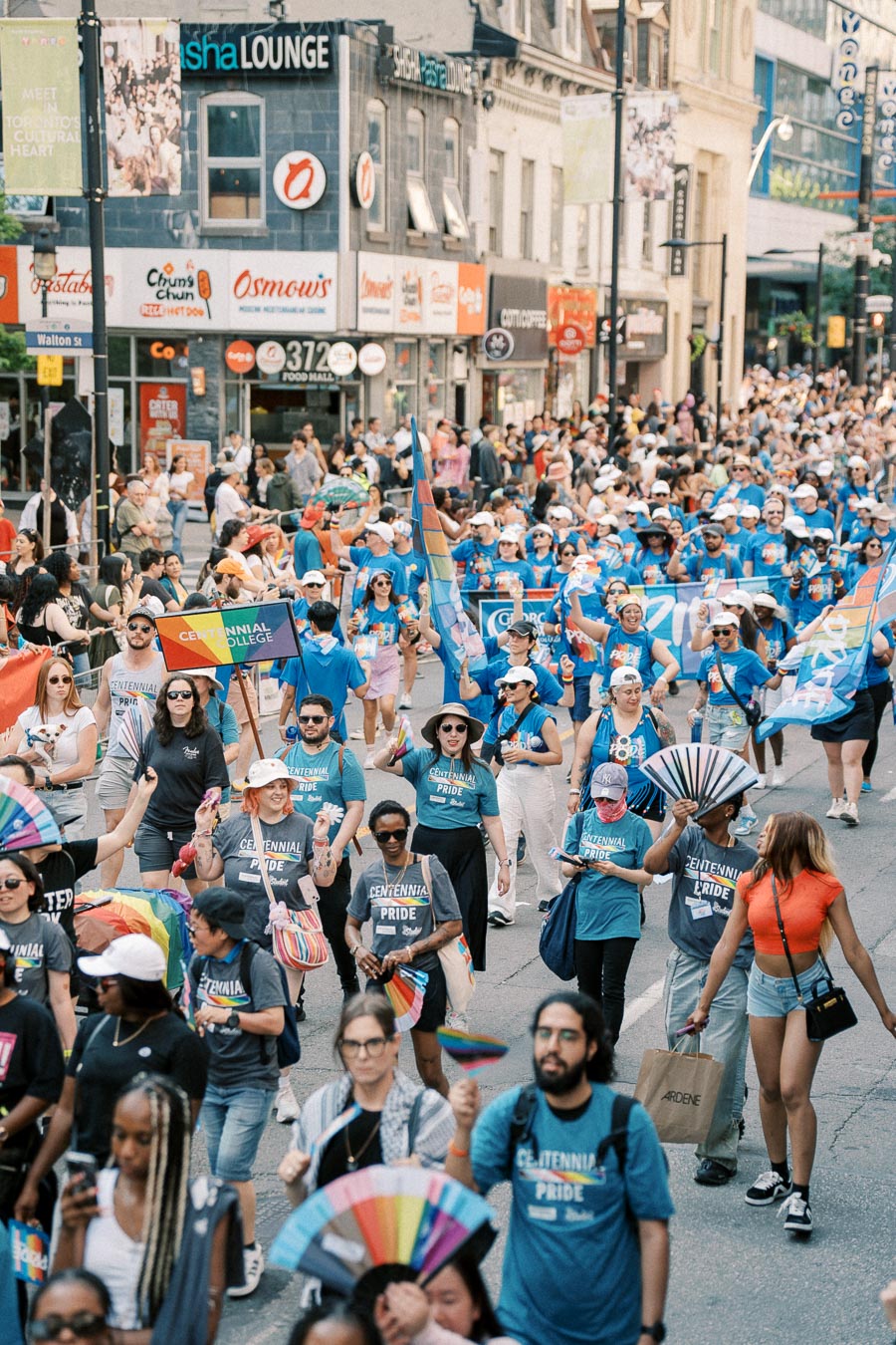 Participants from Centennial College march in a Pride parade, showcasing vibrant rainbow flags and fans. The street is bustling with onlookers and lined with various shops.