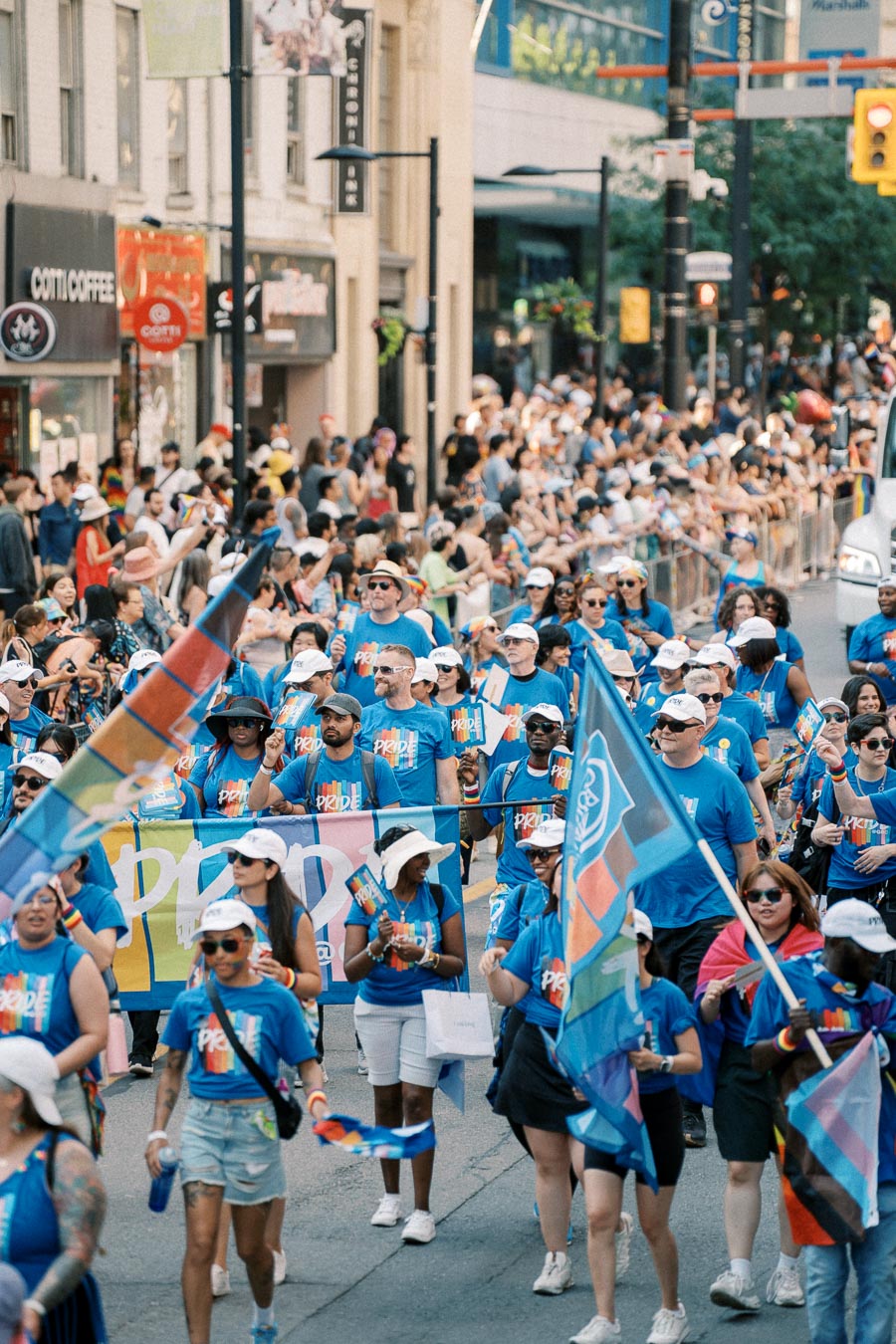 Participants in a Pride parade march down a city street, wearing blue shirts and carrying rainbow flags, with a crowd of spectators lining the sidewalks.