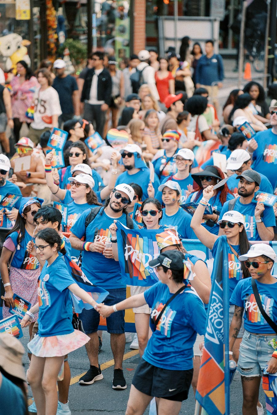 Participants in a Pride parade wearing blue shirts and white caps, celebrating with rainbow flags and banners.